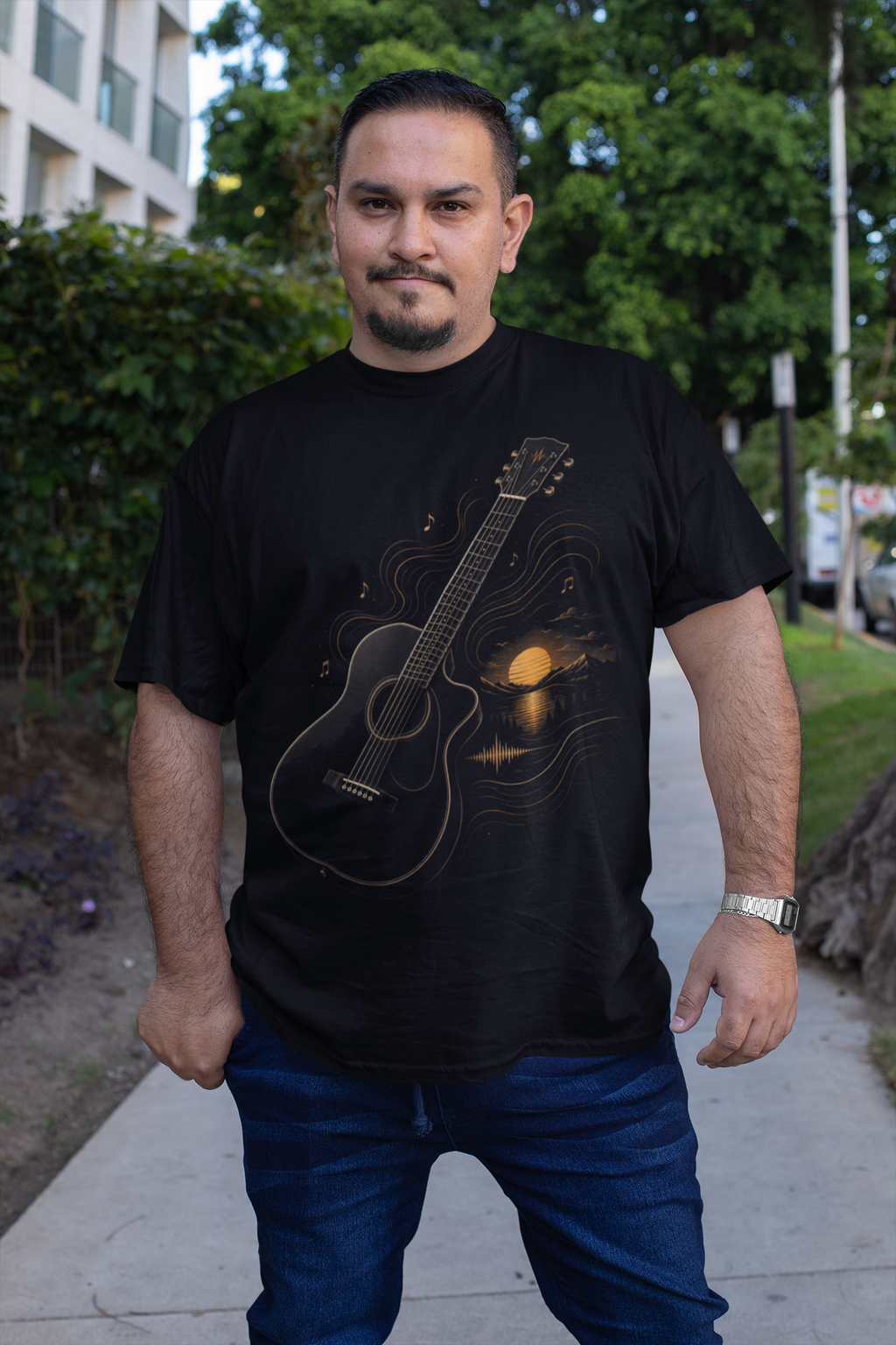 A man wearing a black graphic t-shirt with a guitar and sunset design, paired with blue jeans, standing outdoors on a sidewalk.