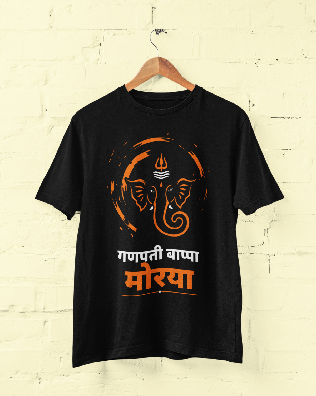 A black short-sleeve t-shirt hanging on a wooden hanger against a light beige painted brick wall. The shirt features a graphic print of a Ganesha head in orange and white, with the Hindi text 'गणपती बाप्पा मोर्या' in white and orange below it.