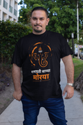 A man wearing a black t-shirt with an orange and white Ganesha graphic and Hindi text, standing outdoors on a sidewalk.