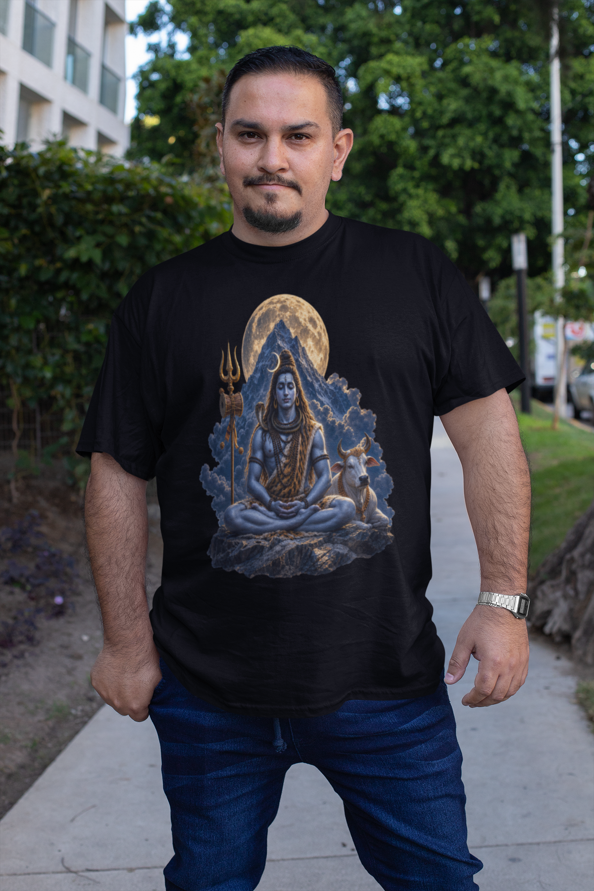 A man wearing a black t-shirt with a detailed graphic of Lord Shiva meditating in front of a full moon, standing on an outdoor sidewalk.